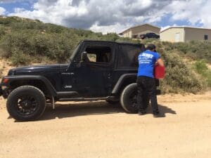 A roadside assistance technician wearing a blue Hoist Towing shirt pouring gasoline from a red container into a black Jeep Wrangler to provide fuel delivery services.