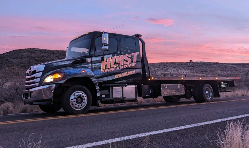 Hoist Towing & Roadside Assistance flatbed tow truck parked street side under a bright pink sunset horizon