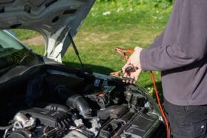 A view under the open hood of a car showing a person connecting booster cables to perform vehicle jump starts next to a grassy area.